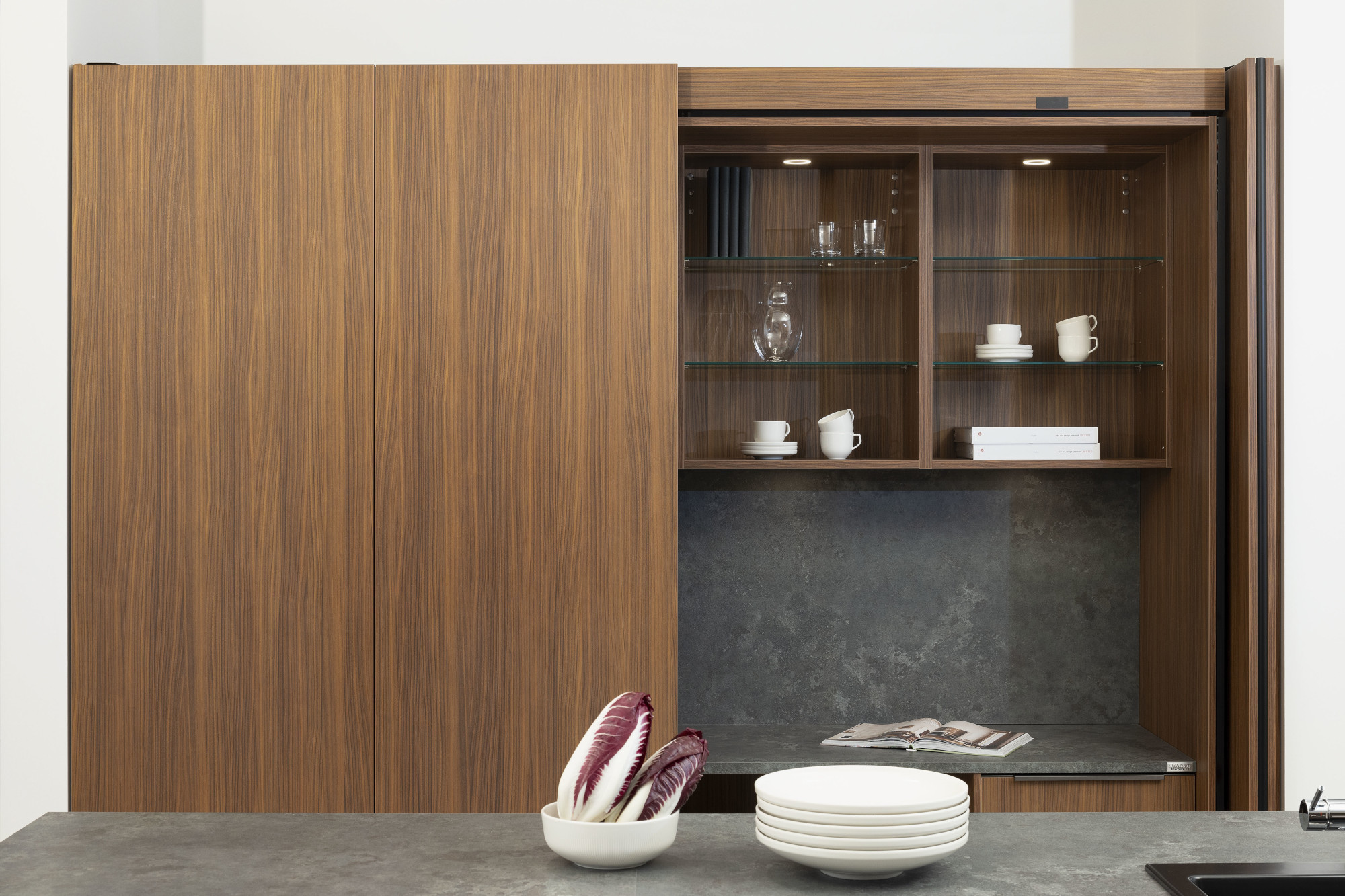 Holzschrank mit Glastüren, darin Geschirr; davor Arbeitsplatte mit Tellern und Chicorée.